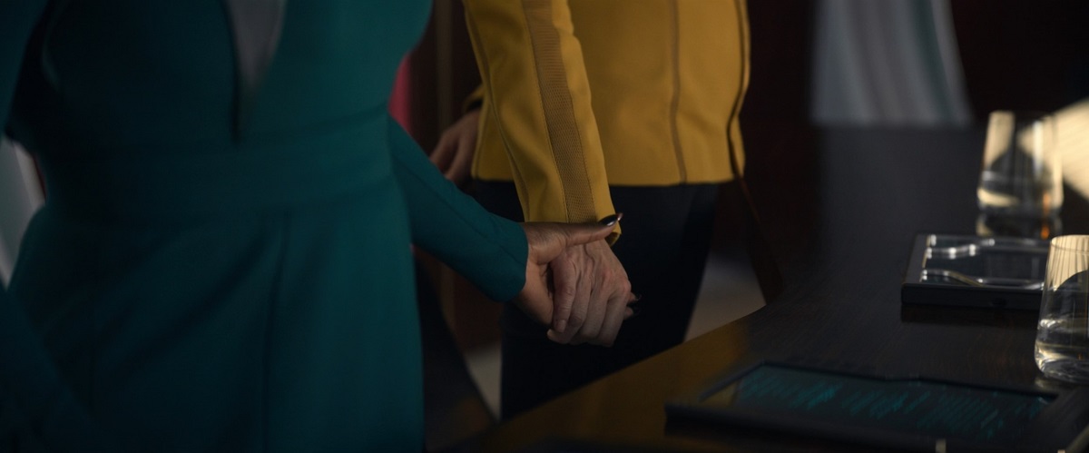 Una (Rebecca Romijn) and Neera (Yetide Badaki) in a scene from 'Strange New Worlds' on Paramount+. We see a close-up of their hands as they hold each other's hand. One a Black woman's who's wearing a blue-green dress. The other a white woman's hand wearing a gold, long-sleeved shirt and black pants as they stand together behind a desk.