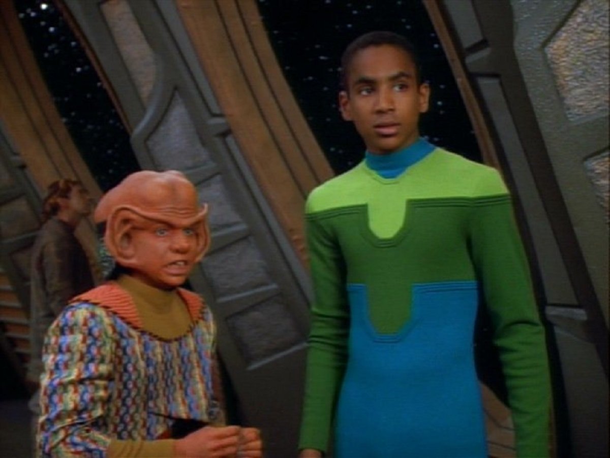 Aron Eisenberg as Nog and Cirroc Lofton as Jake Sisko in a scene from 'Star Trek: Deep Space Nine.' They stand next to each other looking at something warily. Nog is a short Ferengi teenage boy wearing a green, blue, and brown patterned turtleneck sweater. Jake is a taller, Black human teen boy wearing a spandex green and blue bodysuit. 