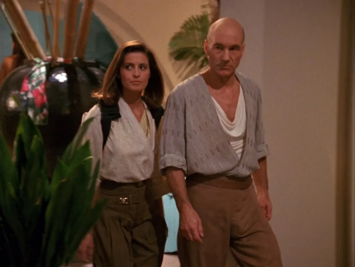 Jennifer Hetrick as Vash and Patrick Stewart as Jean-Luc Picard in a scene from 'Star Trek: The Next Generation.' They are walking side by side as they look off to the side. Vash is a white woman with shoulder-length brown hair wearing a white, long-sleeved shirt and greenish, high-waisted pants. Picard is a bald white man wearing white shirt with a draped neckline under an open-front three-quarter sleeve shirt tucked into high-waisted brown pants. 