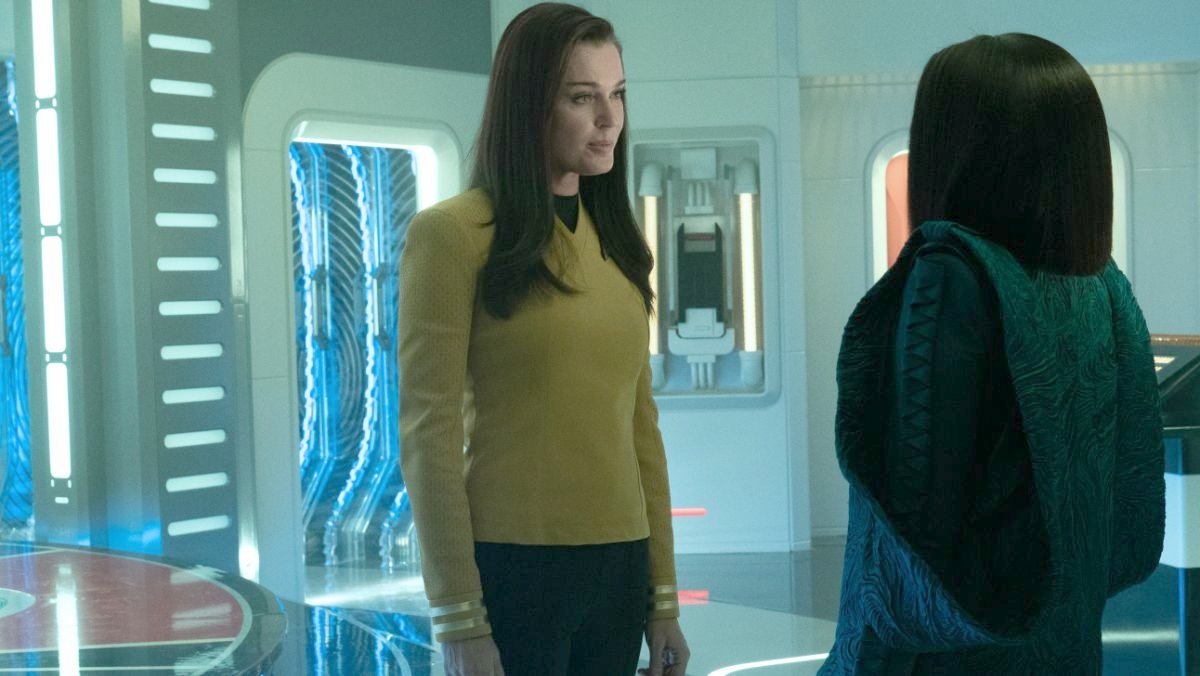Una (Rebecca Romijn) and Neera (Yetide Badaki) in a scene from 'Strange New Worlds' on Paramount+. Una is a white woman with long, dark hair wearing a gold Starfleet uniform. She is standing facing Neera with an annoyed look on her face. Neera is a dark-skinned Black woman with straight black hair wearing a blue-green dress with a blue-green cloak over it with large arm holes. We see her from behind as she faces Una.