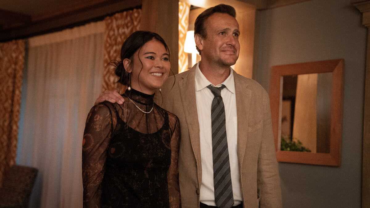 Image of Lukita Maxwell as Alice and Jason Segel as Jimmy in a scene from Apple TV+'s 'Shrinking.' Alice is a mixed race white and Asian teenage girl with her hair in a bun wearing a lacy party dress. Jimmy is a middle-aged white man with short brown hair wearing a beige suit and tie. He has his arm around her shoulder in their home as she smiles and he looks hopeful.