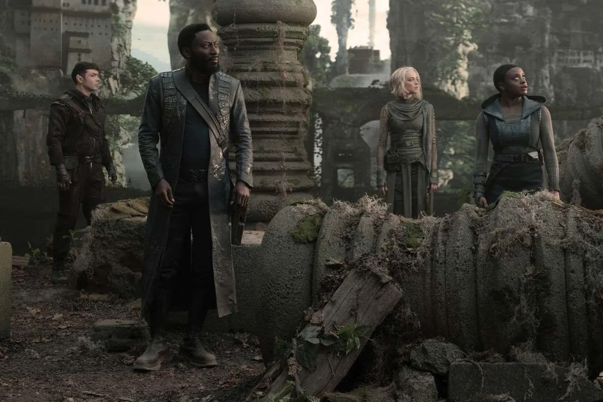 Spock (Ethan Peck), M'Benga (Babs Olusanmokun), Chapel (Jess Bush), and Uhura (Celia Rose Gooding) in a scene from 'Star Trek: Strange New Worlds' on Paramount+. They are standing around ruins on a planet and wearing civilian clothes as they look up at something in the distance.
