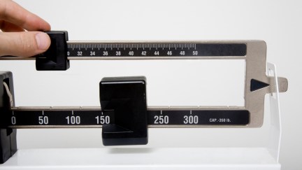 A stand up doctor's office style scale with a hand calculating the weight.