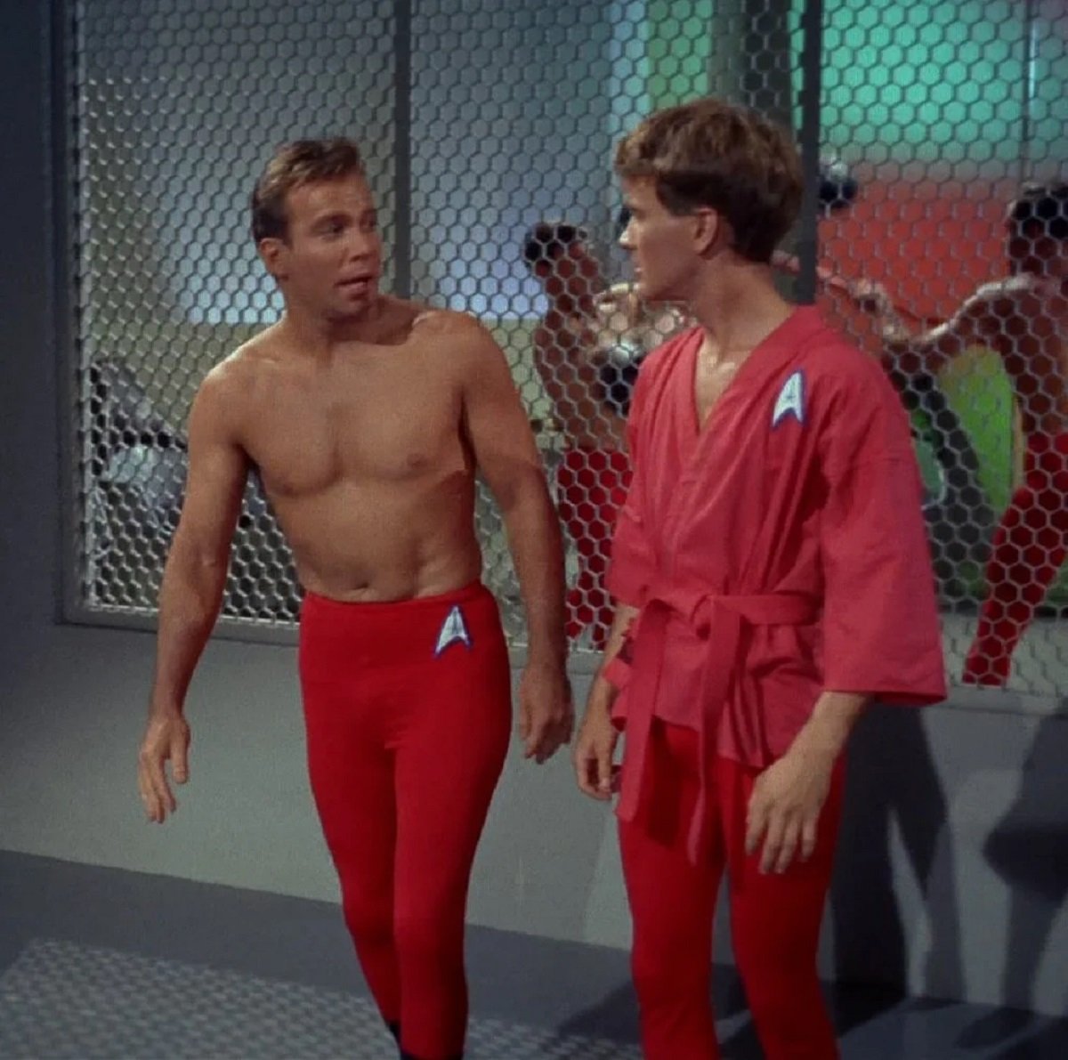 William Shatner as Captain Kirk in a scene from 'Star Trek.' Kirk (white man with short brown hair) is shirtless and wearing tight, red leggings with a Starfleet insignia over one hip. He's standing next to another white man in a red, Starfleet gi with an insignia on the shoulder and the same red leggings. 