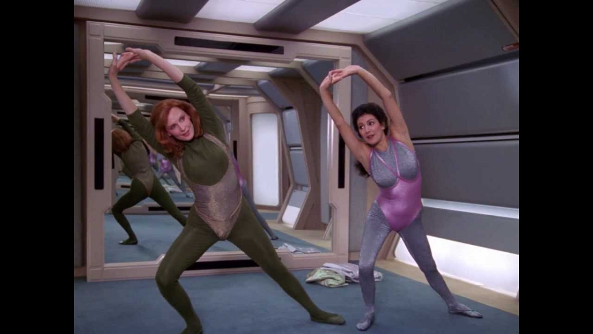 Gates McFadden as Beverly Crusher and Marina Sirtis as Deanna Troi in a scene from 'Star Trek: The Next Generation.' They are lunging and stretching over their heads as they exercise in really tacky exercise outfits. 