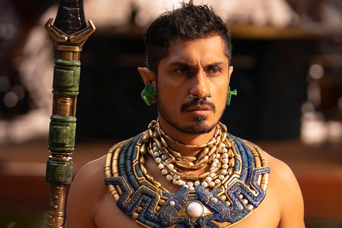 Image of Tenoch Huerta as Namor in Marvel's 'Black Panther: Wakanda Forever.' He is an Indigenous Mexican man with short dark hair with pointy ears and a thin beard. He's wearing green earrings, several beated necklaces and a large blue and gold neck adornment, and he's holding a green and gold spear.