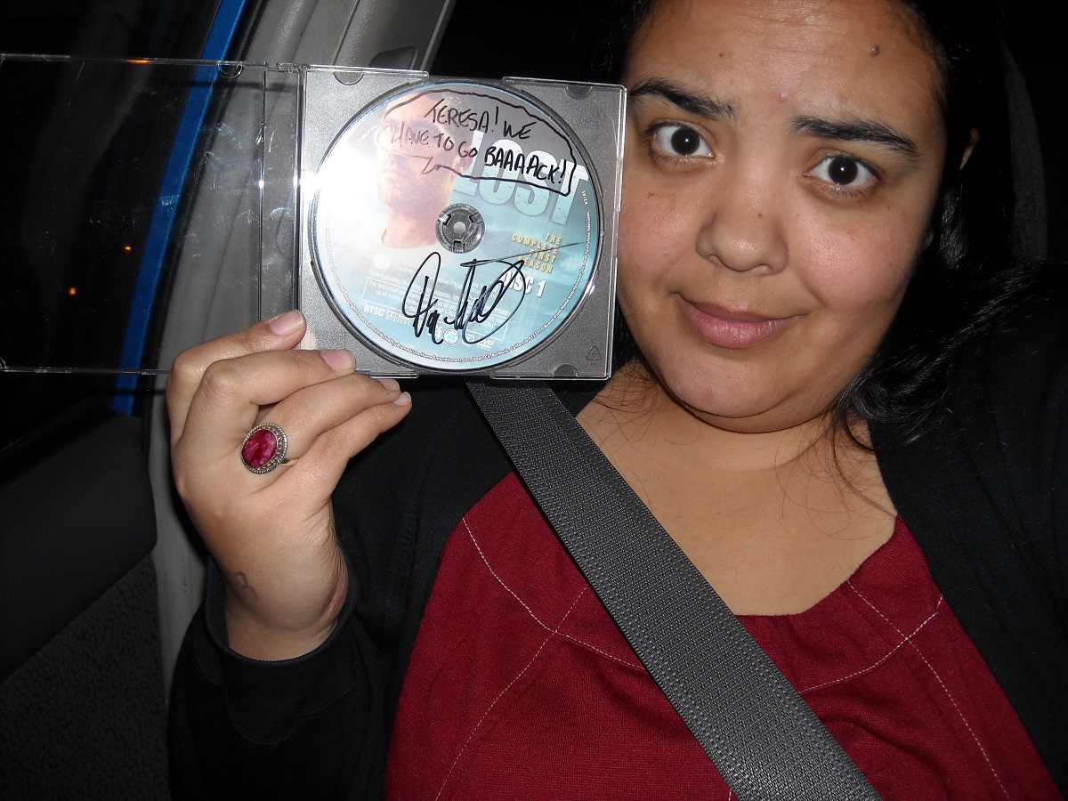 Image of Teresa Jusino (a brown Latina with long dark hair wearing a black cardigan and a dark red dress) sitting in the passenger side of a car holding up a 'Lost' DVD autographed by Damon Lindelof. Lindelof drew a word bubble coming out of Jack's mouth, and it reads "Teresa! We have to go baaaack!"