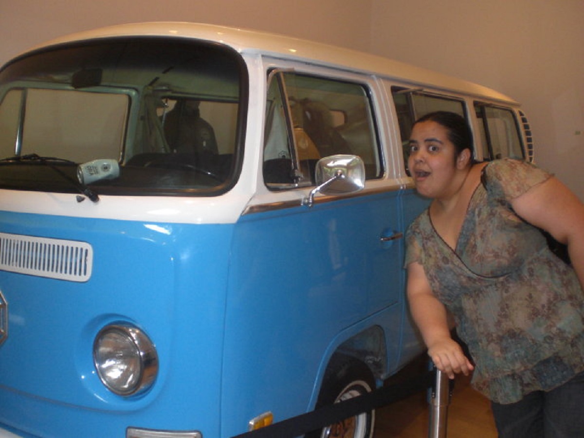 Image of Teresa Jusino (brown Latina with dark hair in a ponytail wearing a green floral shirt) leaning against a light blue and white VW bus from the show 'Lost' at the 'Lost' Exhibit in New York in 2010.
