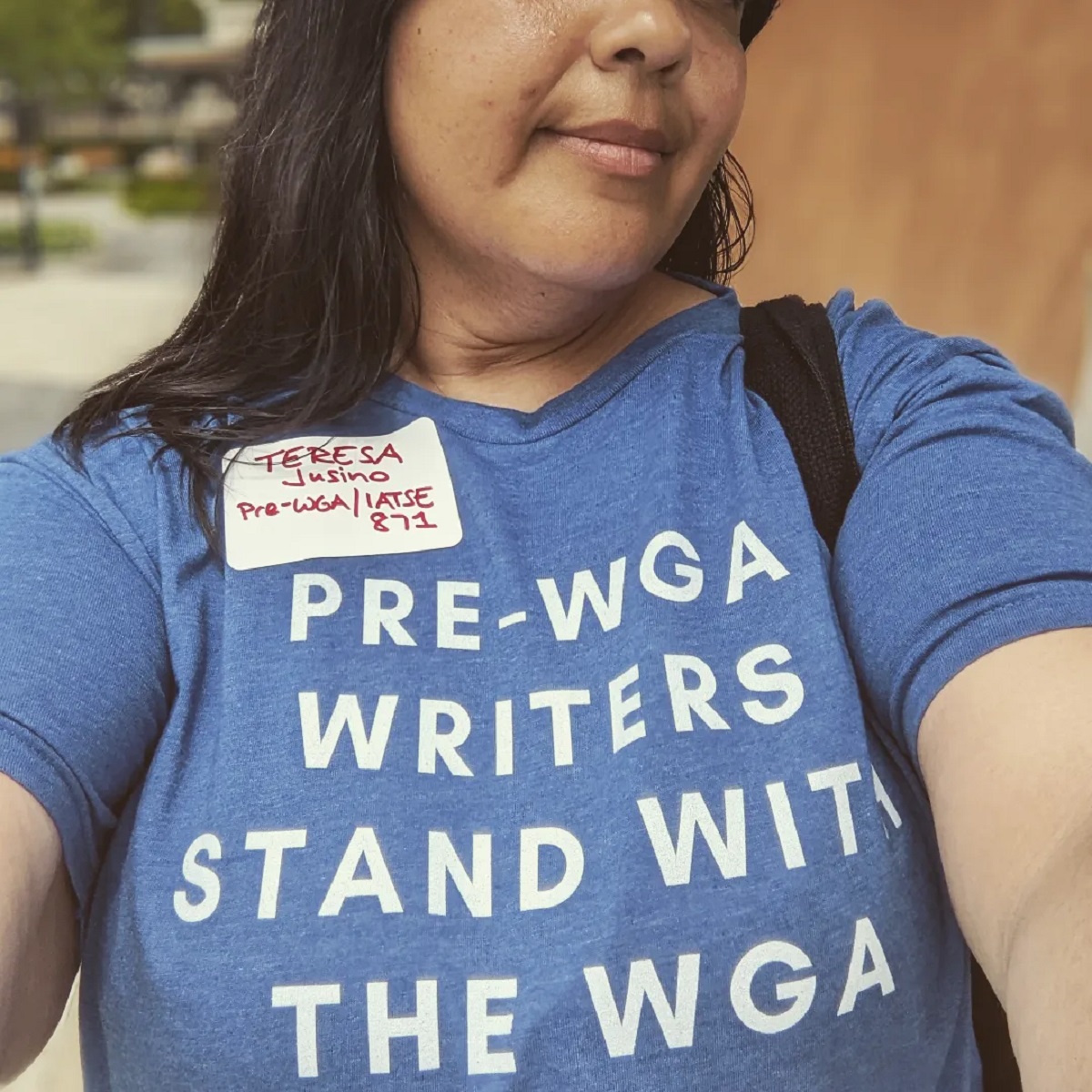 Square image of Teresa Jusino (a brown Latina with long, black hair) from the nose down showing off her t-shirt. The t-shirt is blue and reads "Pre-WGA Writers Stand With the WGA." She's also wearing a nametag that reads "Teresa Jusino - Pre-WGA/IATSE 871.