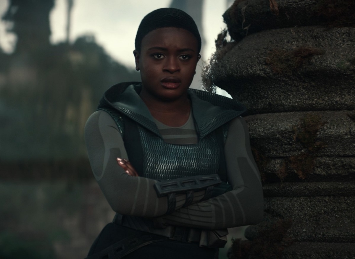 Uhura (Celia Rose Gooding) in a scene from 'Star Trek: Strange New Worlds.' She is a young Black woman with close-cropped short black hair. She's leaning against a stone pillar with her arms folded as she watches someone speak. She's wearing a blue, hooded pullover vest over a tight, long-sleeved dark/light grey shirt. 