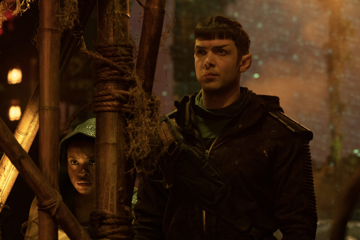 Ethan Peck as Spock and Celia Rose Gooding as Uhura in a scene from 'Star Trek: Strange New Worlds.' They are hiding behind a wooden structure trying to see something going on in the room. Spock is a white Vulcan with short, Black hair wearing a black jacket and a green shirt. Uhura is standing slightly behind him with her blue hood up. 