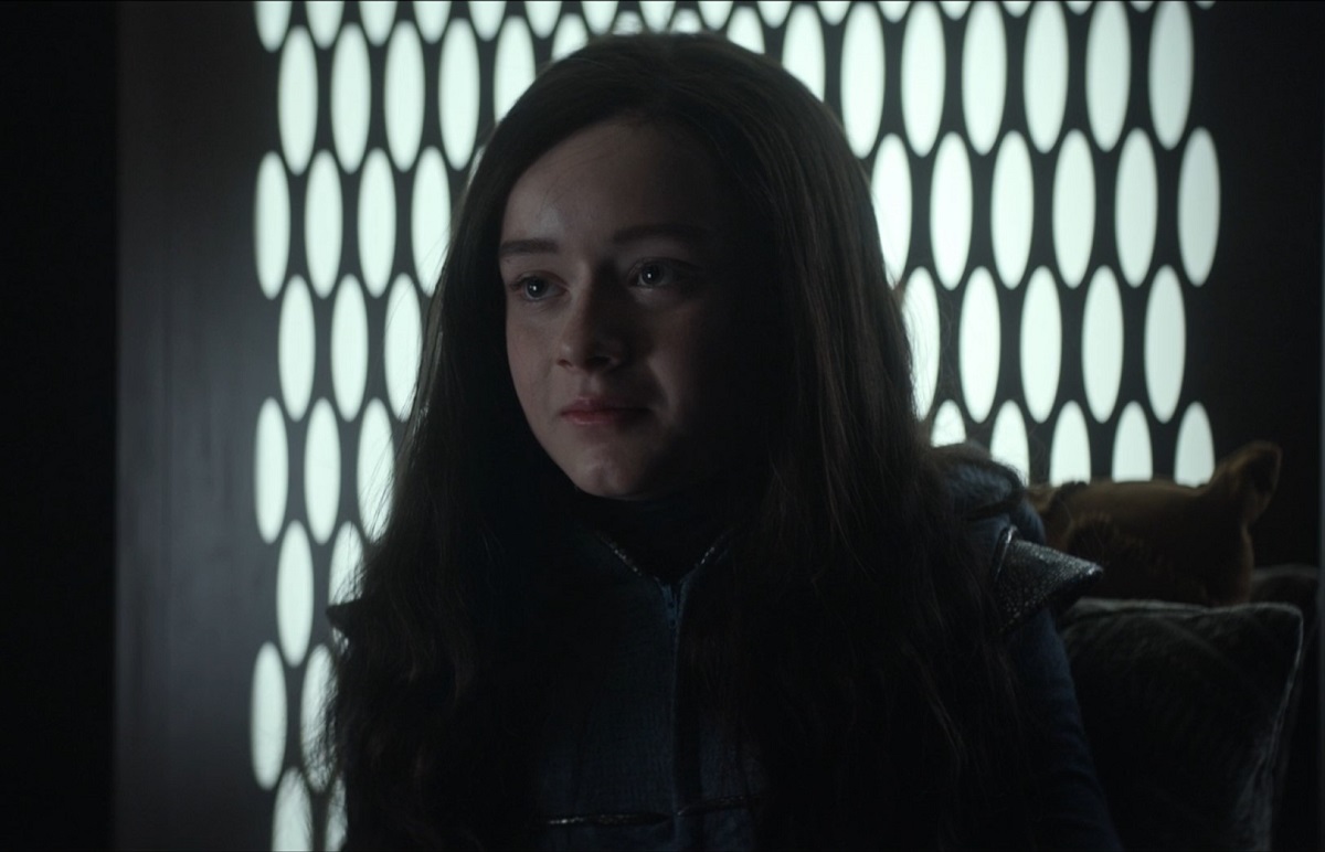 Young Una (Anna Claire Beitel) in a scene from 'Strange New Worlds' on Paramount+. Young Una is a white "tween" girl with long, black hair wearing a blue jumpsuit that zips up the front. We see her from the shoulders up as she looks at someone off camera with a serious expression.