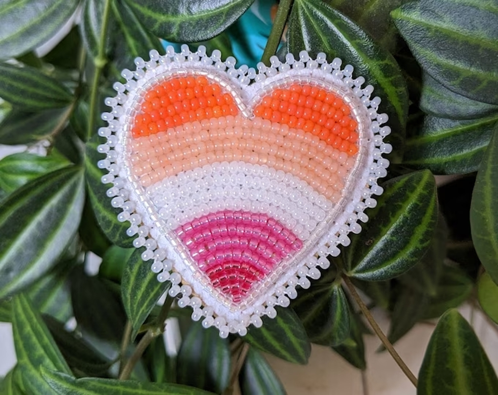 A beaded lesbian flag heart on a background of leaves, created by anim0shbeads