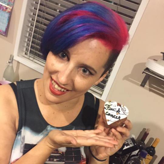 A young woman (Brittany Knupper) with bright pink and blue hair holds a cupcake with icing reading "fuck cancer"