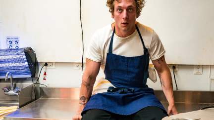 Jeremy Allen White as Carmy in 'The Bear': A white man with a frazzled look on his face wears a blue apron and sits on a metal kitchen table.