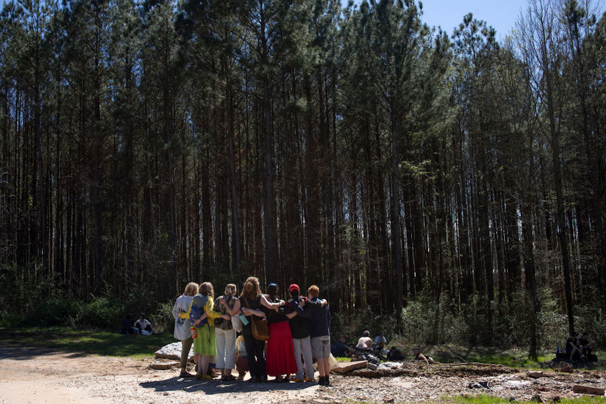 A group of about a dozen people embrace, with the tall trees of a forest behind them.