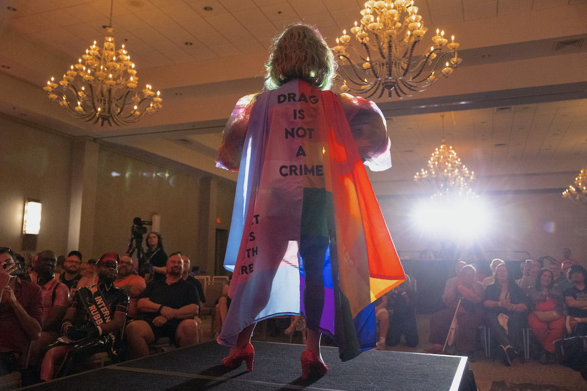 A drag performer is seen on a catwalk from behind, wearing a rainbow cape that reads "Drag is not a crime."