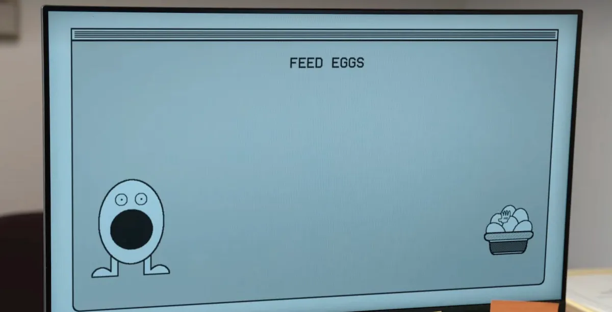 A computer screen with a black and white game on it. At the top, the game says "FEED EGG." On the left is an egg with a face and its mouth wide open. On the right is a basket of smaller eggs.