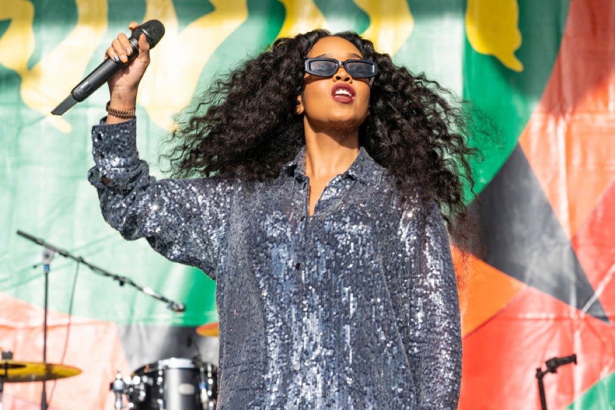 Gabriella Wilson, known professionally as H.E.R., performs during 2023 New Orleans Jazz Heritage Festival. She's holding a microphone above her head.