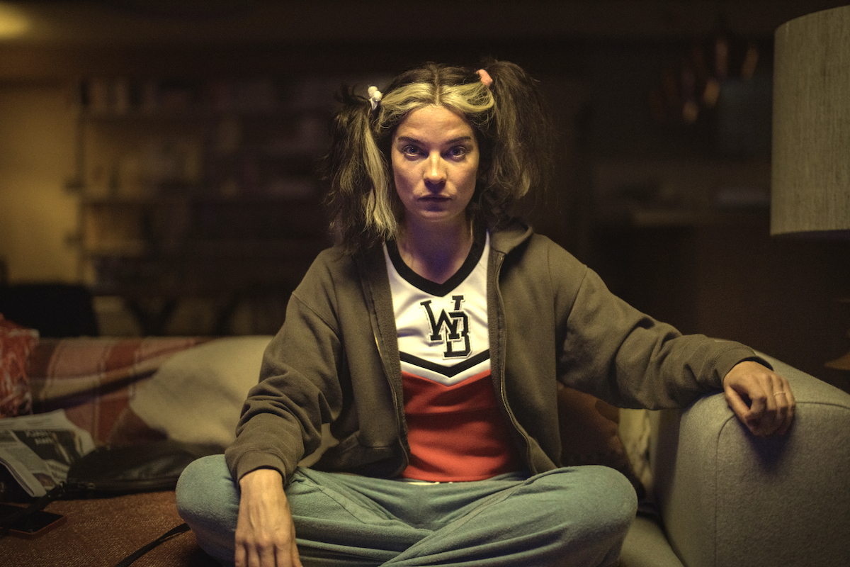Joan (Annie Murphy) sits on the couch in a cheerleading costume, with her hair in messy pigtails.