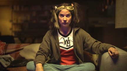 Joan (Annie Murphy) sits on the couch in a cheerleading costume, with her hair in messy pigtails.