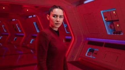 La'an stands in a red hallway on the Enterprise, looking at something off camera.