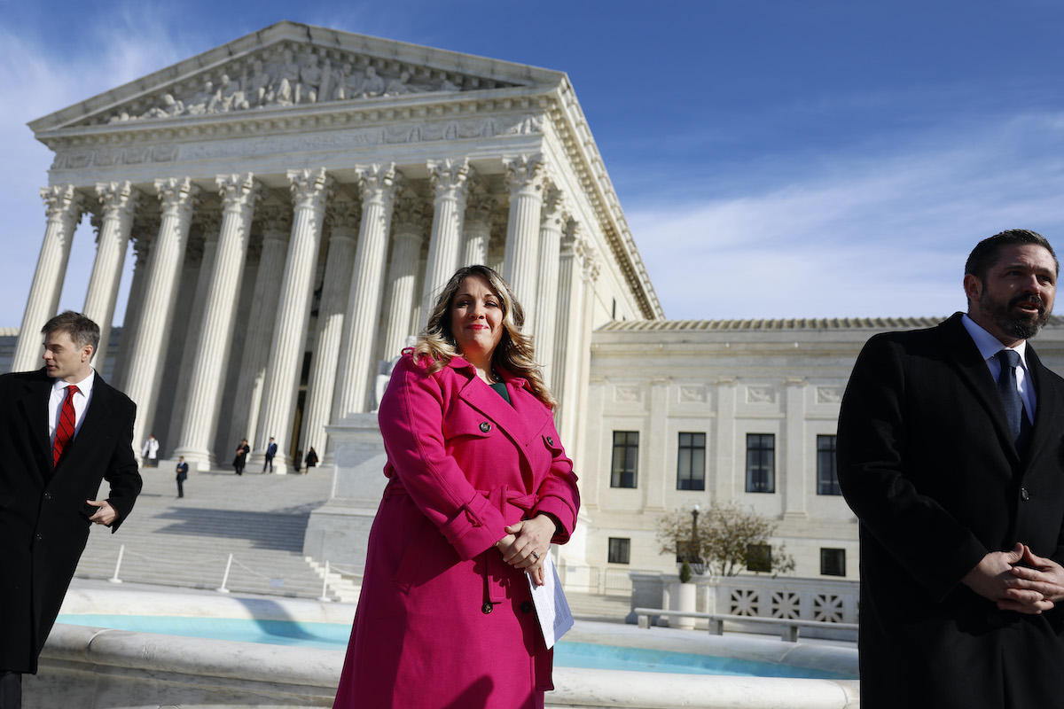 A white woman in a pink blazer stands in front of the Supreme Court building and smirks at the camera.