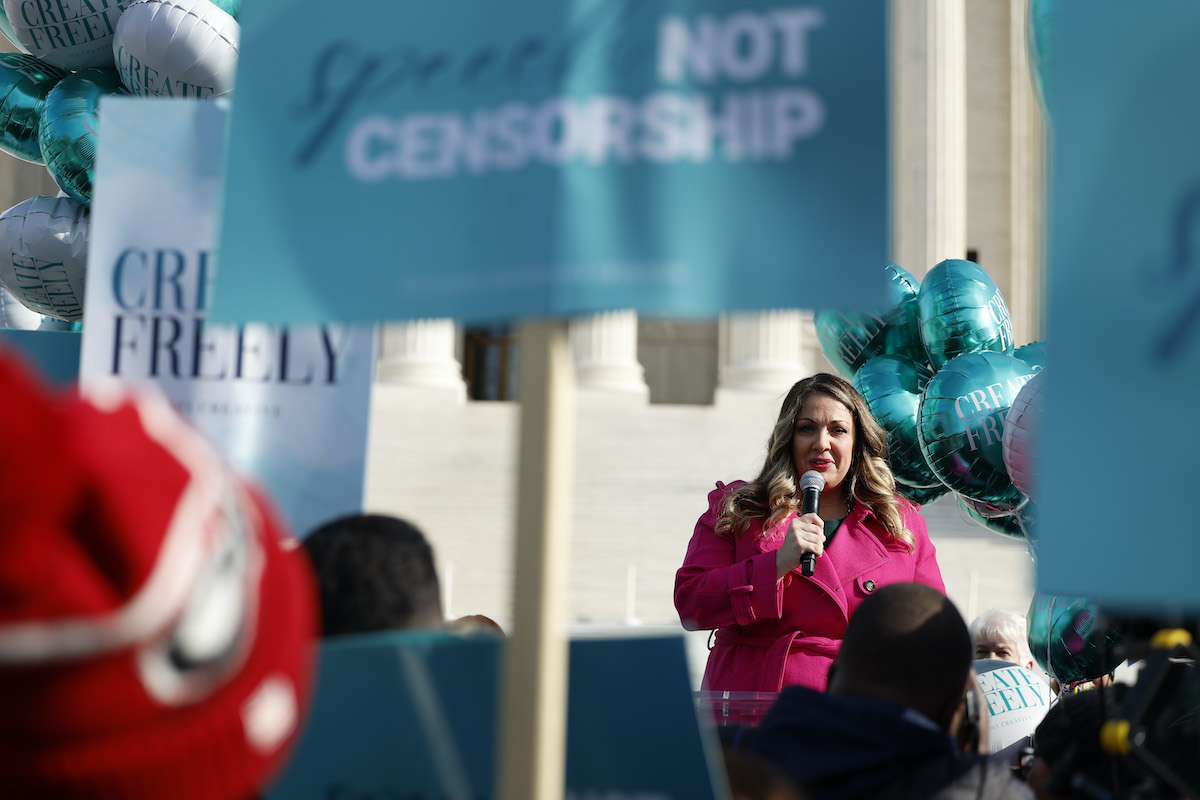 A white woman (Lorie Smith) in a bright pink blazer speaks into a microphone during a rally outside the Supreme Court.