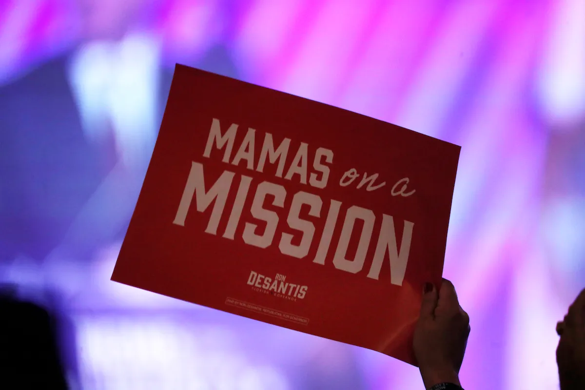 A woman's hand holding a sign reading "Mamas on a mission," with a DeSantis campaign logo beneath.
