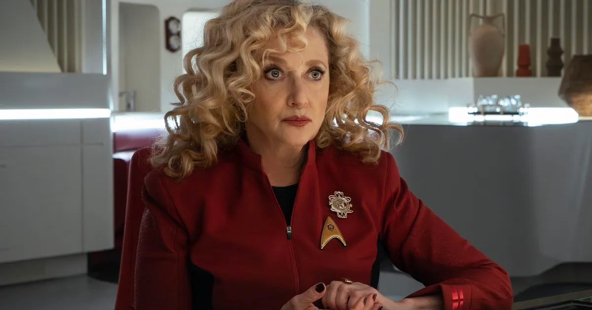 Pelia (Carole Kane) sits at a table in a white room, wearing a Starfleet uniform.