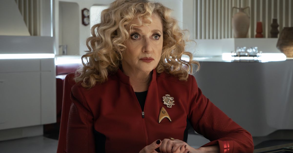 Pelia (Carole Kane) sits at a table in a white room, wearing a Starfleet uniform.