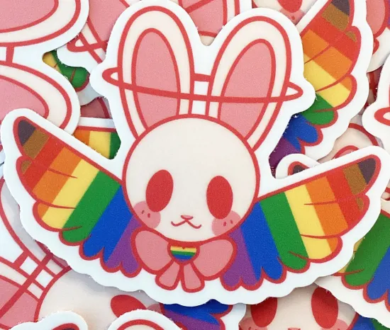 A sticker featuring a pink cartoon line drawing of a rabbit's face with rainbow angel wings an a pink bow tie.