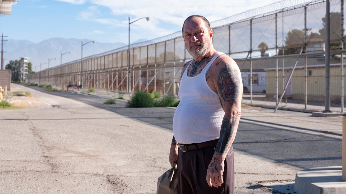 Fuches, wearing a wifebeater and numerous tattoos, glares at someone outside of a prison yard.