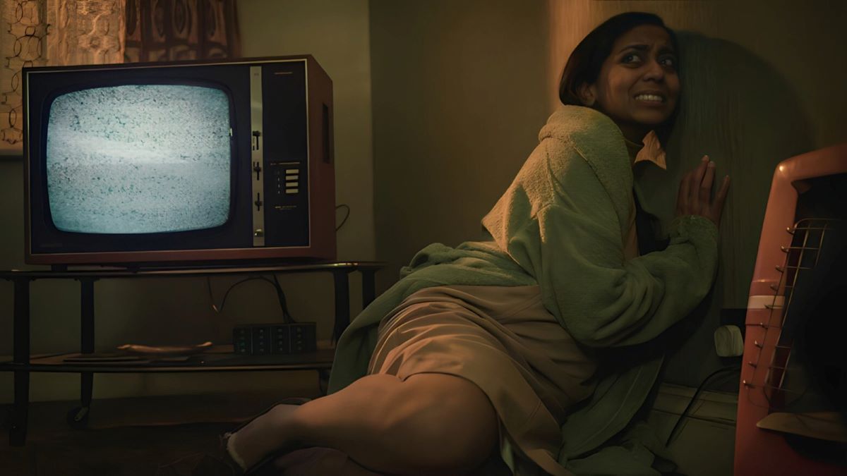 A woman cowers in the corner of her living room, with a TV showing static next to her.