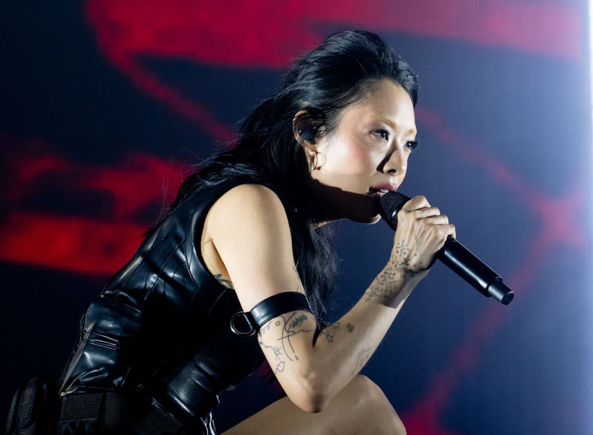 Rina Sawayama leans over and belts into a microphone, wearing black leather.