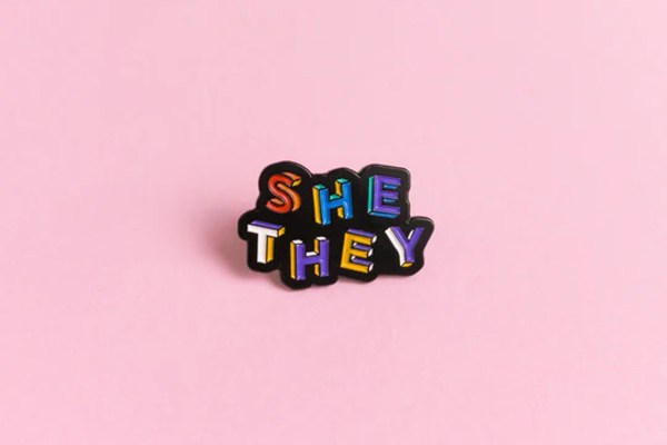 Heckin' Unicorn She/They enamel pin badge: She is on the line above they, the letters are in rainbow colours.