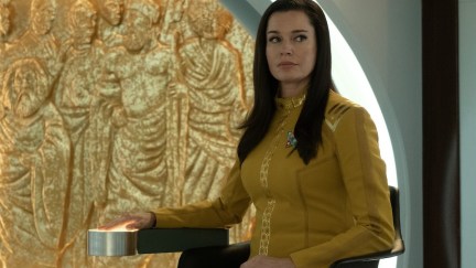 Una (Rebecca Romjin) sits in front of a gold sculpture showing a crowd of people. Her hand is over a glowing pad, and she watches someone off camera.