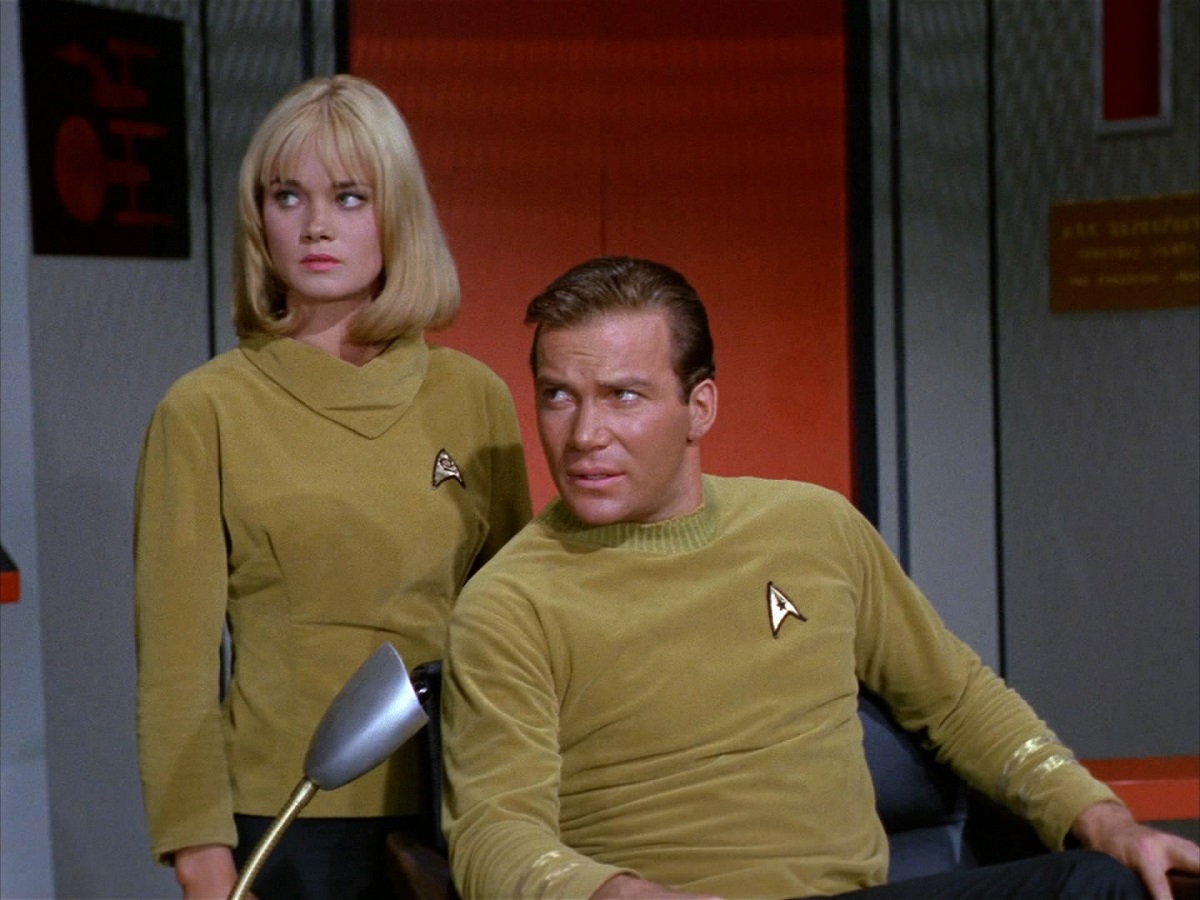 Yeoman Smith (Andrea Dromm) and Captain Kirk (William Shatner) in a scene from 'Star Trek.' Smith is a blonde woman with a neck-length bob with bangs. She's wearing a gold Starfleet turtleneck and standing behind Kirk, a white man with brown hair wearing a gold Starfleet shirt and sitting in his captain's chair. They're both looking off camera.
