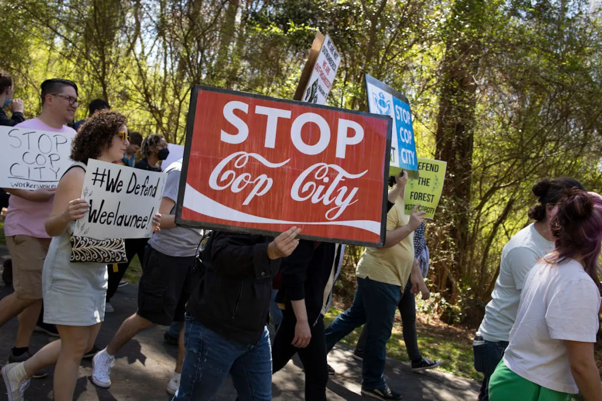 Protesters carry signs through a forest, including one that says "Stop Cop City" in a Coca Cola font.