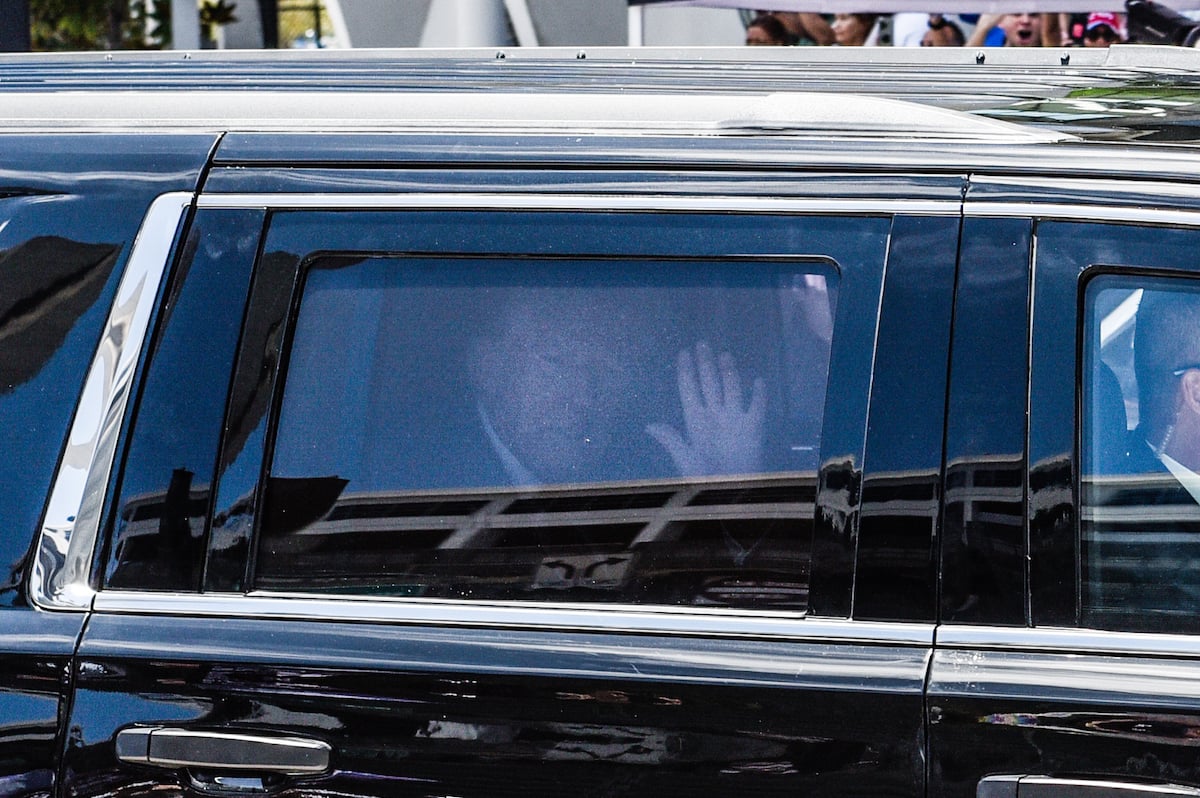 Donald Trump sits in the backseat of a car, waving through a tinted window.