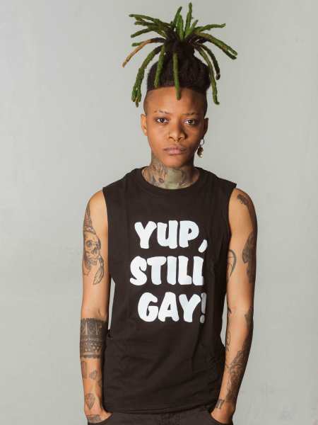 A Black woman with locs in a high ponytail wears a blank sleeveless shirt with "yup, still gay" written on it in thick white letters.