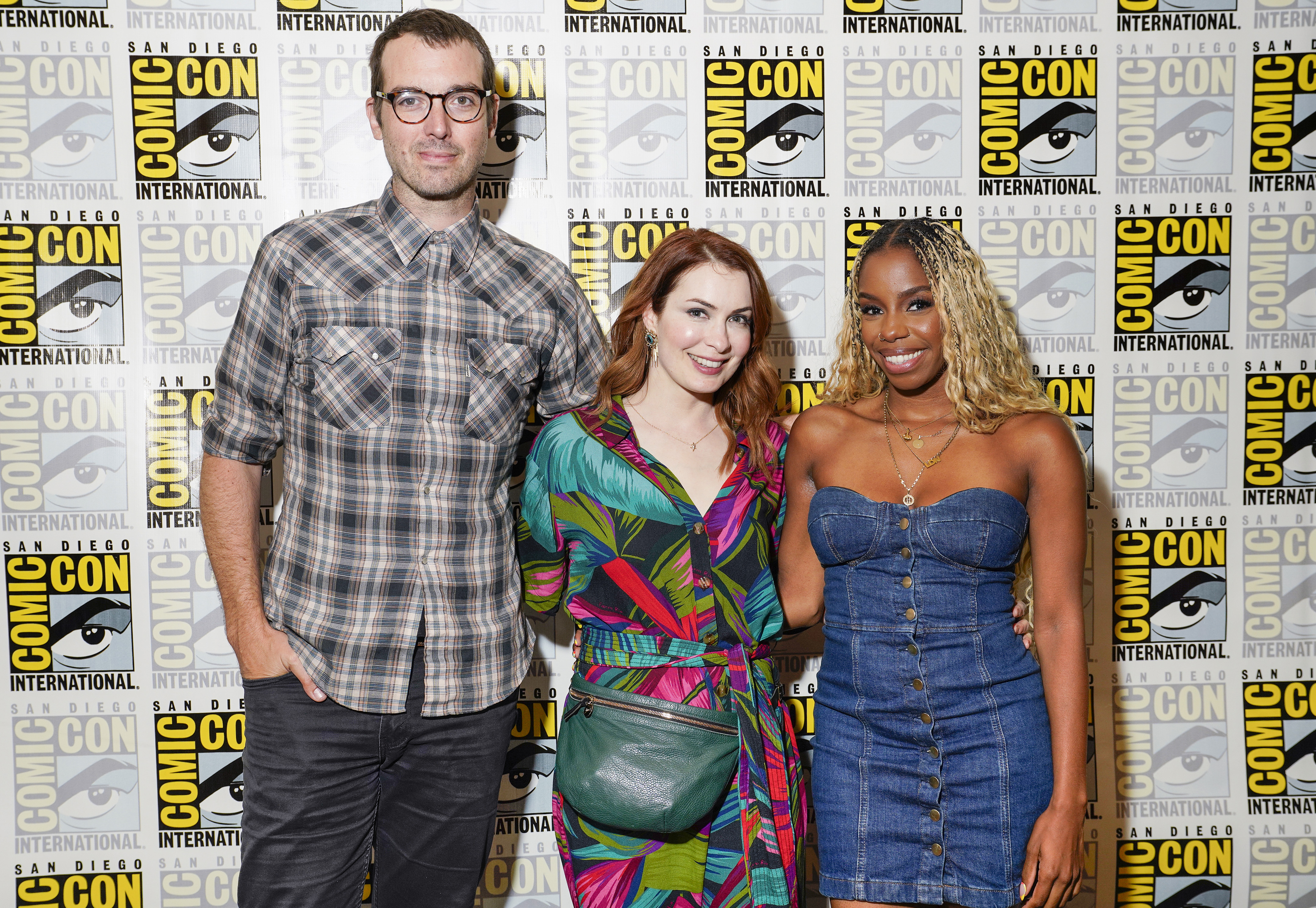 Jonah Ray Rodrigues (tall, white man with short dark hair and glasses wearing a brown plaid buttondown and black pants), Felicia Day (white woman with long red hair wearing a blue/green/pink/black plant print jumpsuit and a green purse), and London Hughes (Black woman with long, curly blonde hair with black roots wearing a denim strapless dress) stand smiling in front of a Comic-Con step and repeat. 