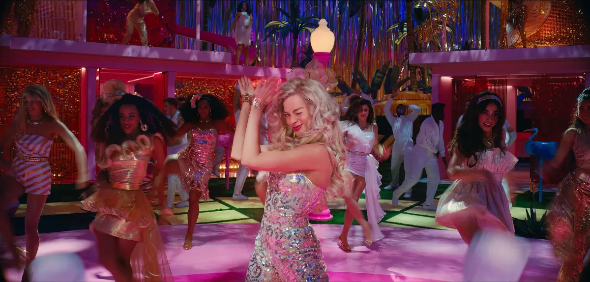 Margot Robbie's Barbie dancing, holding up her hands and clapping while winking, in the midst of a Barbie dance party in Greta Gerwig's Barbie movie.