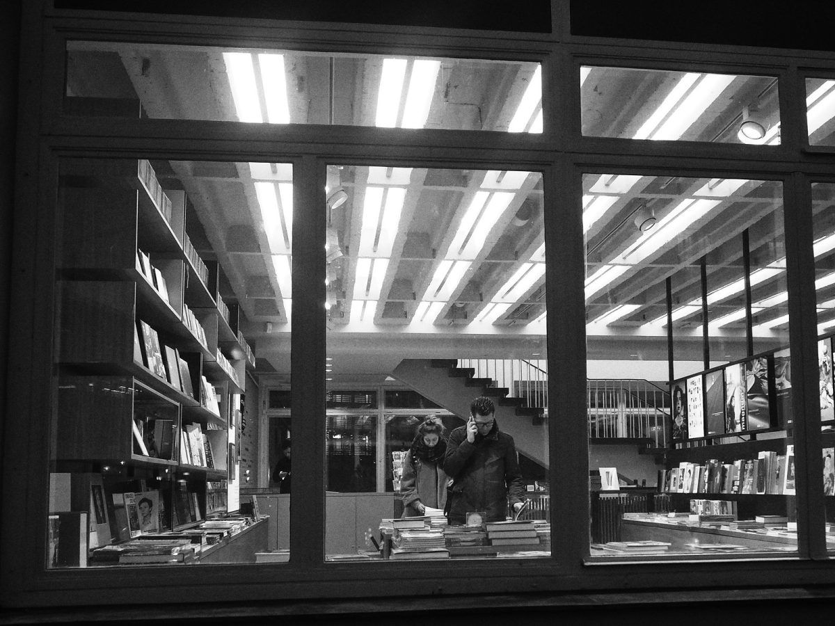Black and white image showing the inside of a bookstore through clear windows. Two people are visible in the photo looking down a table full of books.