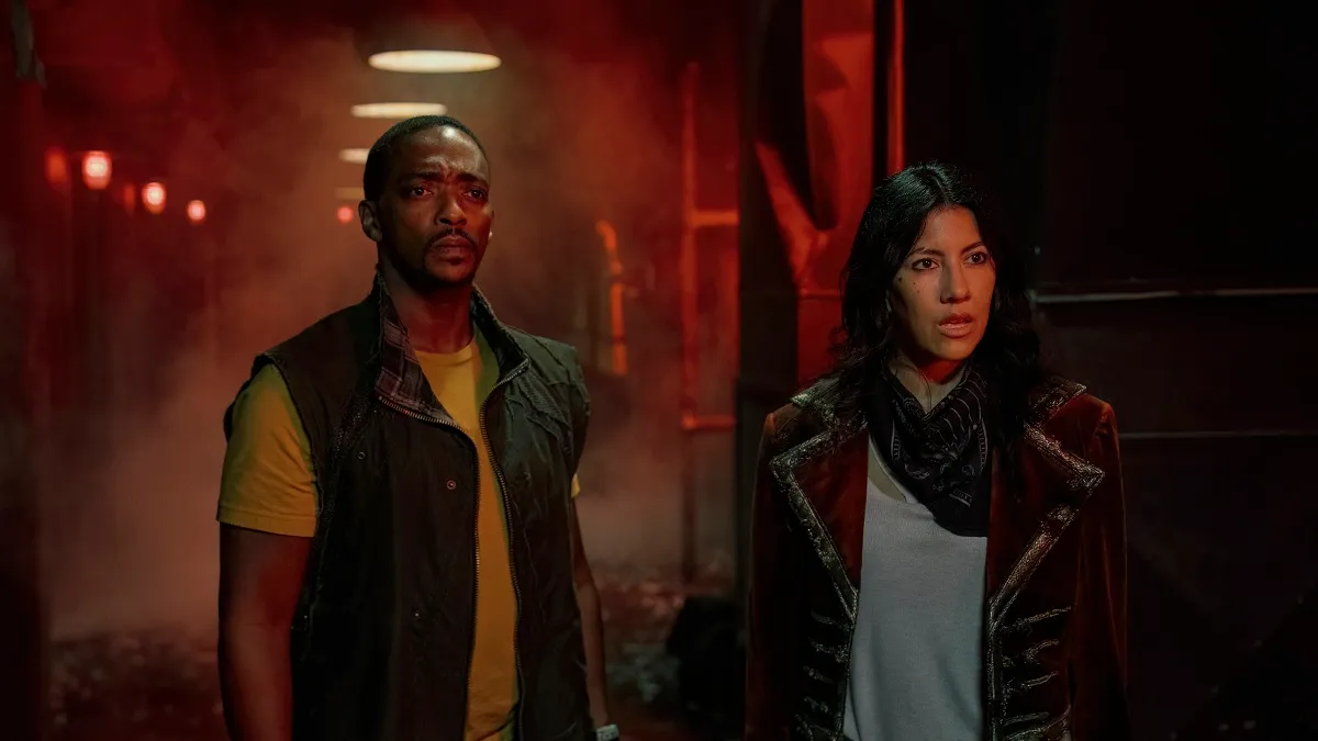 Anthony Mackie (Black man with close-cropped hair, thin beard, wearing a yellow t-shirt under a green zippered vest) as John Doe Stephanie Beatriz (brown Latina with shoulder-length dark hair wearing a black scarf and grey shirt under a dark maroon blazer) as Quiet in a scene from Peacock's 'Twisted Metal.' they are standing side-by-side in a dark industrial hallway looking toward something that seems unpleasant.
