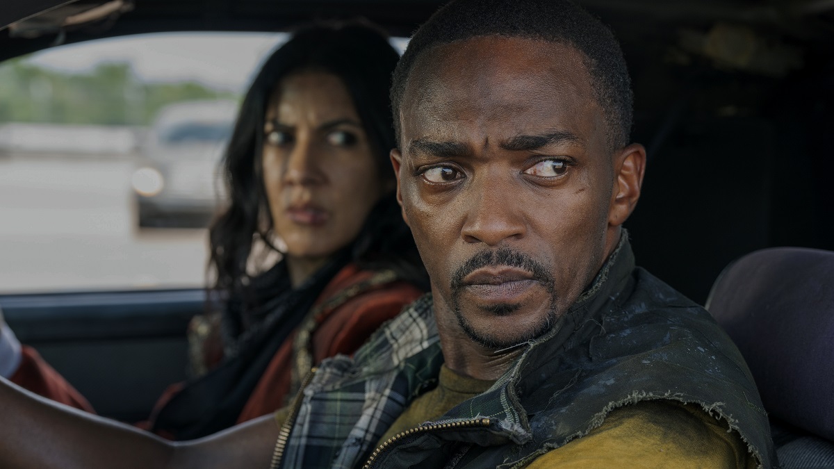 Stephanie Beatriz (brown Latina with shoulder-length dark hair wearing a black scarf and grey shirt under a dark maroon blazer) as Quiet, and Anthony Mackie (Black man with close-cropped hair, thin beard, wearing a yellow t-shirt under a zippered vest) as John Doe in a scene from Peacock's 'Twisted Metal.'  John is in the driver's seat of a car with Quiet as a passenger. They are both looking at something weird and concerning out the window. John is the foreground and Quiet is out of focus. 