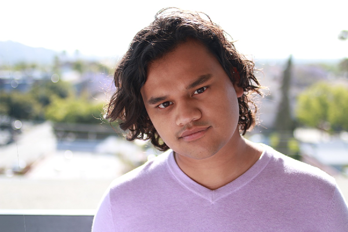 Headshot of film director Bishal Dutta. Dutta is a young, Indian man with brown skin and chin-length, wavy, dark hair. He is wearing a greyish-pink v-neck shirt as he stands outside on a balcony. 