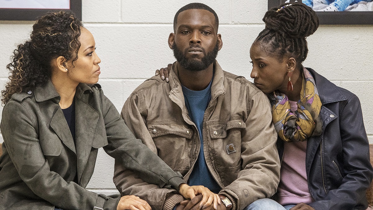 Dawn-Lyen Gardner as Charley, Kofi Siriboe as Ralph-Angel, and Rutina Wesley as Nova in a scene from OWN's 'Queen Sugar.' They are all Black and seated in a waiting room. The two women are comforting their brother in the middle, who is clearly upset. Charley has long, curly, dark hair and is wearing a grey coat. Ralph-Angel has closely cropped dark hair and a beard and wears a brown jacket and a blue t-shirt, and Nova has her dark, braided hair pulled up on top of her head and wears a black leather jacket, a yellow patterned scarf, and a pink t-shirt.