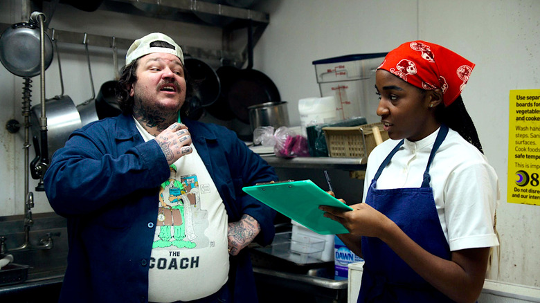 A white man and a Black woman holding a clipboard have a conversation in a restaurant kitchen.