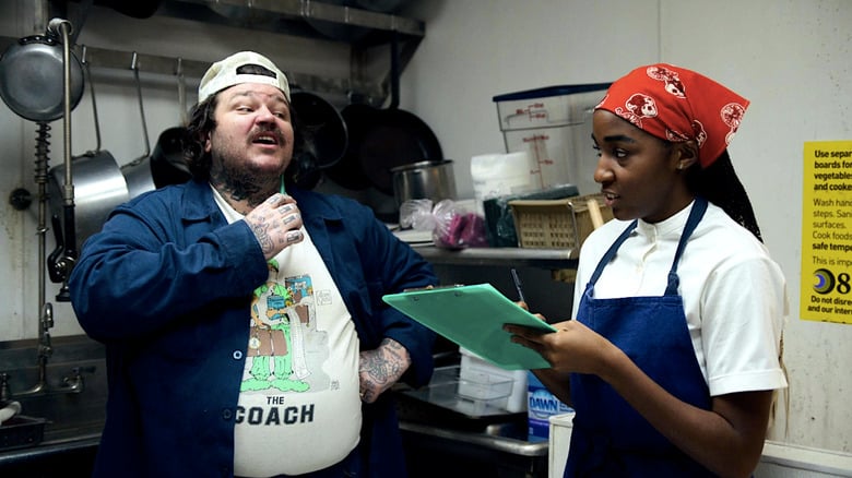 A white man and a Black woman holding a clipboard have a conversation in a restaurant kitchen.